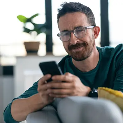 Man with glasses and mustache wearing a teal shirt looking at his phone while sitting on a sofa in a modern home interior
