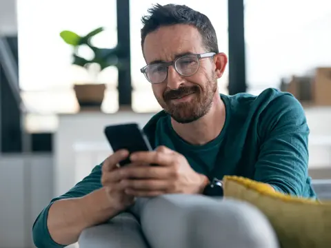 Man with glasses and mustache wearing a teal shirt looking at his phone while sitting on a sofa in a modern home interior