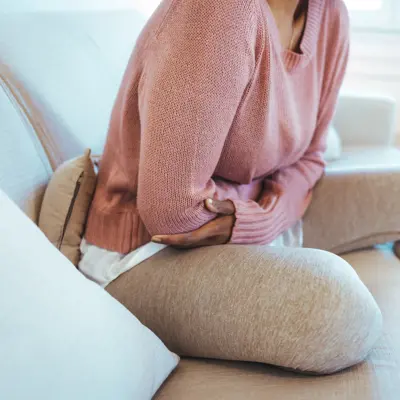 Woman in pink sweater sitting on a white couch holding her stomach with both hands, suggesting abdominal discomfort or pain