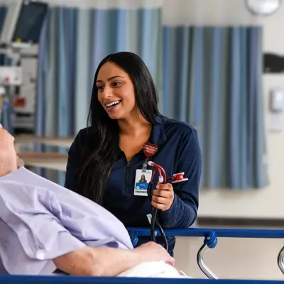 Female health care team member in navy scrubs smiling while speaking with a male patient lying in a hospital bed in a surgical services unit