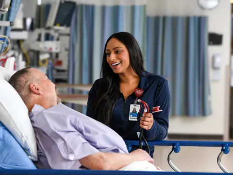 Female health care team member in navy scrubs smiling while speaking with a male patient lying in a hospital bed in a surgical services unit