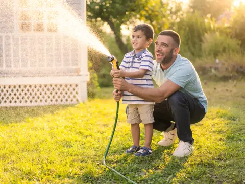 Father kneeling behind young son in striped shirt, both holding garden hose together and spraying water across sunlit lawn in front of white house