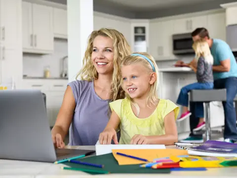A mother and daughter are working on homework together at a kitchen table, with other family members in the background.