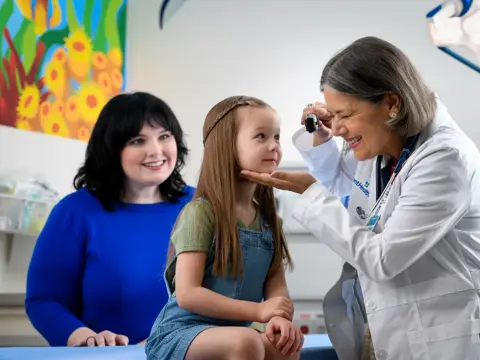 Doctor examining a young girl's eyes with a woman standing nearby in a clinic.