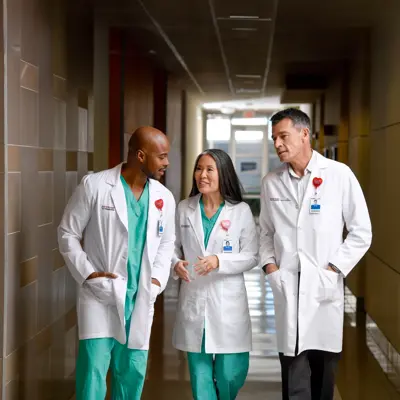 Three healthcare professionals in white coats and green scrubs walking down a hospital corridor.