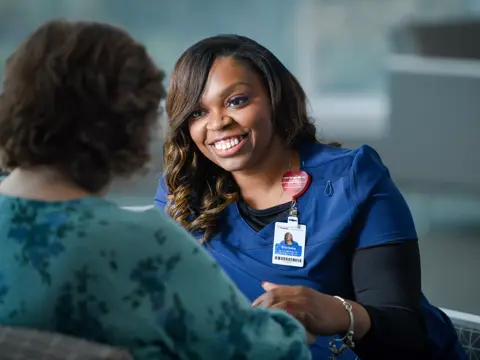 A smiling nurse in blue scrubs talks to a patient, conveying warmth and care.