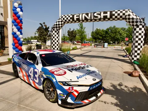 AdventHealth and UChicago Medicine branded NASCAR race car number 43 positioned beneath a checkered finish line arch at an outdoor event with patriotic balloon decorations
