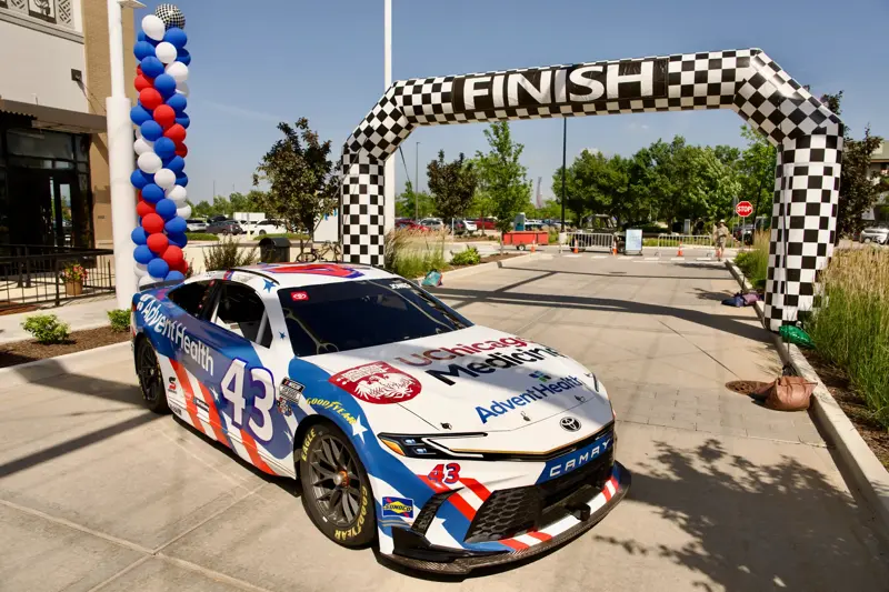 AdventHealth and UChicago Medicine branded NASCAR race car number 43 positioned beneath a checkered finish line arch at an outdoor event with patriotic balloon decorations