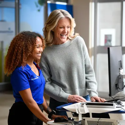 A smiling woman in a blue AdventHealth shirt assists an older woman at a computer station.