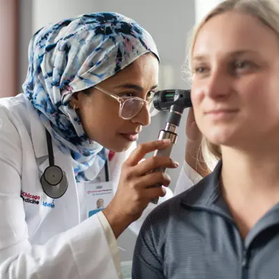 A doctor wearing a white coat and a scarf examines a patient's ear with an otoscope.