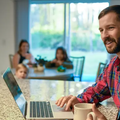 Man in plaid shirt working on laptop at home with family in background.