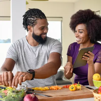 A man and woman in a kitchen preparing a salad together, smiling and using a tablet.