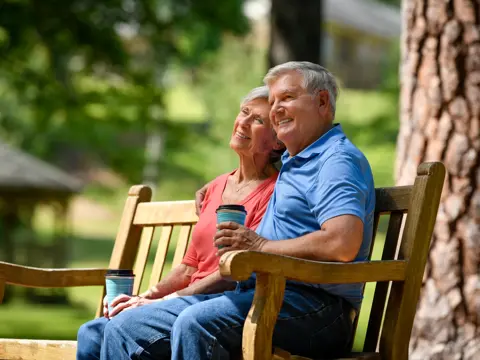 Smiling senior couple sitting together on a wooden park bench, holding coffee cups and enjoying a peaceful moment outdoors in a sunny garden setting