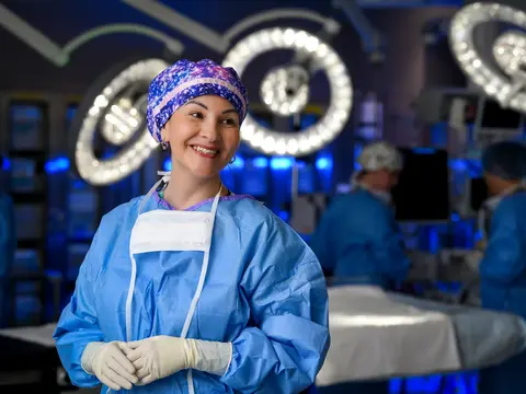 Smiling female surgeon in blue scrubs and patterned surgical cap standing in an operating room with illuminated surgical lights in the background