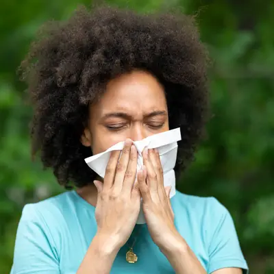 Woman with curly hair in a turquoise shirt blowing her nose into a white tissue while standing outdoors with a blurred green background