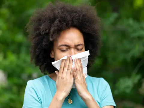 Woman with curly hair in a turquoise shirt blowing her nose into a white tissue while standing outdoors with a blurred green background