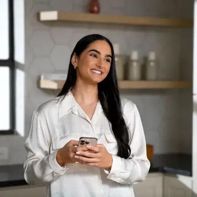 Smiling Hispanic woman in white shirt holding smartphone standing in modern kitchen with wooden shelves and white cabinets