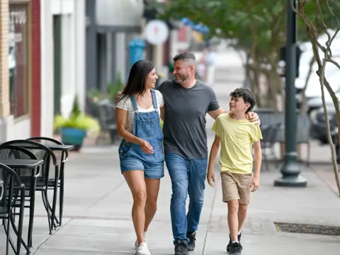 Family of three walking together on a downtown sidewalk, with a father in the middle embracing his teenage daughter and son as they smile and enjoy quality time together