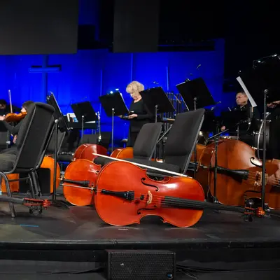 Orchestra performing on stage with cellos and violinists, illuminated by blue lighting.