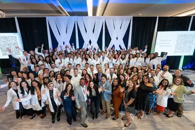 Large group of people, many wearing white coats, smile and wave while posing together on a decorated stage at a white coat ceremony.