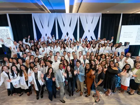 Large group of people, many wearing white coats, smile and wave while posing together on a decorated stage at a white coat ceremony.