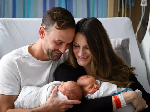 A smiling couple holding their newborn twins in a hospital room.