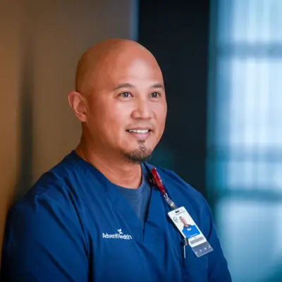A smiling man in blue scrubs with an AdventHealth badge stands against a wall.