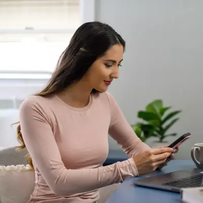A woman in a pink shirt is sitting at a desk, using her phone and laptop with a candle lit.