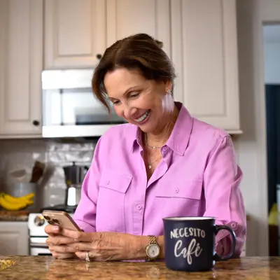 A smiling woman in a pink shirt uses her phone in a modern kitchen with a black mug nearby.