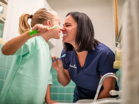 A mother and daughter are brushing their teeth together in a bathroom, smiling and laughing.