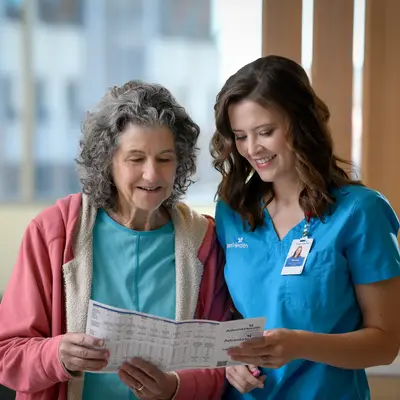 Health care professional in blue scrubs reviewing medical documents with a smiling older female patient wearing a pink cardigan in a bright hospital corridor