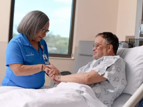 A nurse in a blue uniform holds the hand of a smiling patient in a hospital bed.