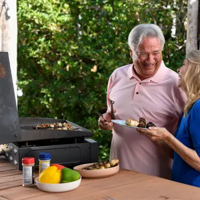 Mature couple smiling and enjoying grilled food together on an outdoor patio with a grill and fresh vegetables on a wooden table