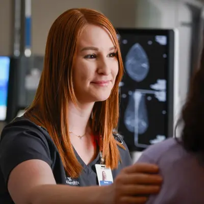 A smiling nurse with red hair consoles a patient in a medical setting with a mammogram on the screen.