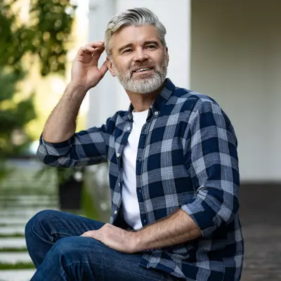 Smiling gray-haired man with beard sits outdoors in plaid shirt and jeans, hand raised to his head.