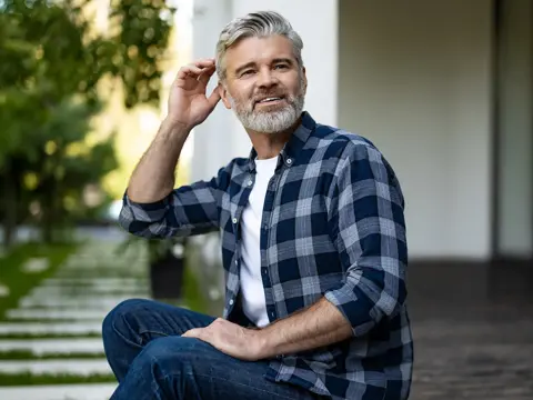 Smiling gray-haired man with beard sits outdoors in plaid shirt and jeans, hand raised to his head.