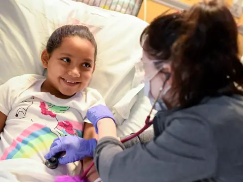 A smiling young girl in a hospital bed as a healthcare professional checks her heart with a stethoscope.