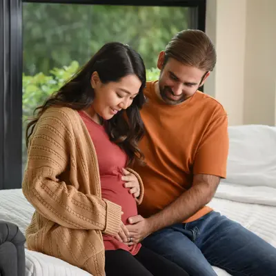 A pregnant woman and her partner sit together on a bed, smiling and holding her belly.