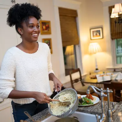 A woman in a white sweater is cooking pasta in a kitchen with a dining table in the background.