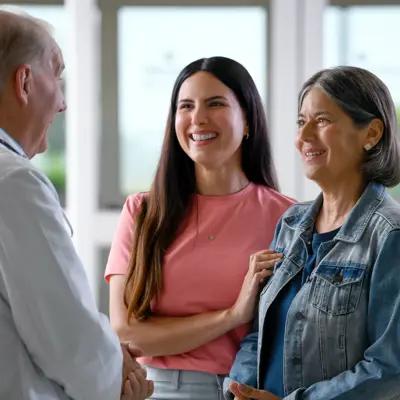 A doctor in a white coat talks with two smiling women, one in a denim jacket, in a bright clinic.