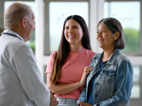 A doctor in a white coat talks with two smiling women, one in a denim jacket, in a bright clinic.