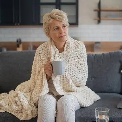 Person with short light hair sits on a sofa wrapped in a cream knit blanket, holding a mug beside an open laptop and a glass of water.
