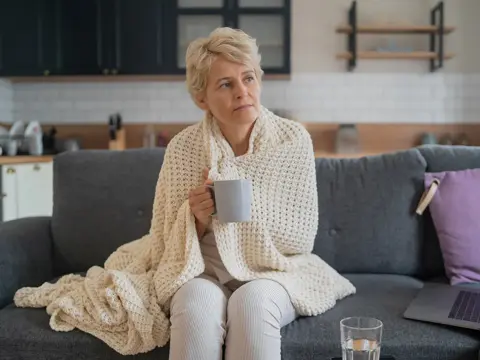 Person with short light hair sits on a sofa wrapped in a cream knit blanket, holding a mug beside an open laptop and a glass of water.