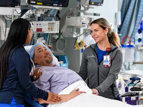 Two AdventHealth team members in scrubs and an AdventHealth jacket attending to a smiling male patient lying in a hospital bed in a medical facility with monitoring equipment visible in the background.