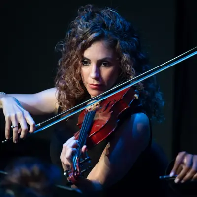 A woman with curly hair plays a red violin in a concert setting, illuminated by stage lights.