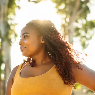 A woman stands outdoors with hands on hips, sunlight behind them, wearing a sleeveless top.