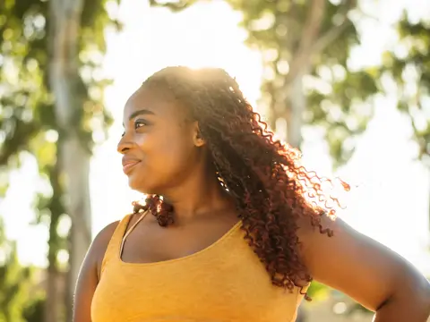 A woman stands outdoors with hands on hips, sunlight behind them, wearing a sleeveless top.