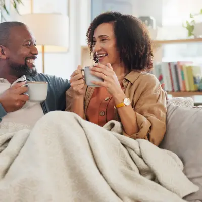 Couple sipping from mugs under a blanket on their sofa