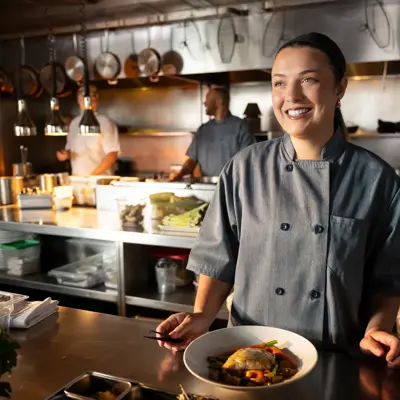 A smiling chef stands behind a counter in a busy kitchen, holding a plate of food.