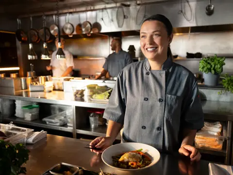 A smiling chef stands behind a counter in a busy kitchen, holding a plate of food.